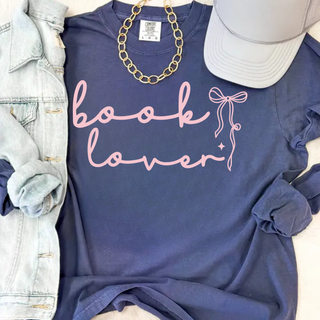Book Lover Coquette Bow Top in Denim Comfort Colors tee with blush pink script lettering and delicate coquette bow accent, flat lay styled with denim jacket, gold chain necklace, and grey trucker hat