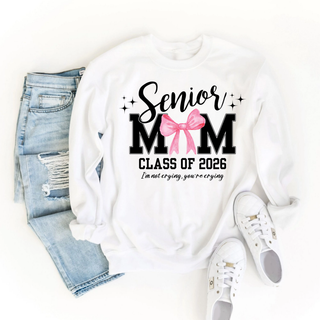 Senior Mom Class of 2026 Graphic Crewneck in white with black and pink text and bow, includes phrase I’m not crying, you’re crying, displayed flat with jeans and white sneakers.