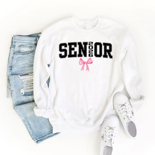 SENIOR 2026 black varsity text with pink bow graphic on white crewneck sweatshirt styled with light wash distressed denim jeans and white sneakers flat lay on white background