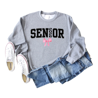SENIOR 2026 black varsity text with pink bow graphic on gray crewneck sweatshirt styled with medium wash denim jeans and white Converse sneakers flat lay on light background