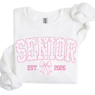 SENIOR pink varsity lettering with bow pattern fill and pink outline, EST. bow graphic 2026 printed on white crewneck sweatshirt with Gildan tag visible at collar