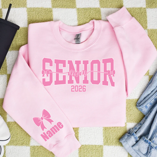 SENIOR mom 2026 pink varsity lettering with bow detail printed on pink crewneck sweatshirt, sleeve graphic showing bow with Name customization text, styled flat lay with denim jeans, black tumbler and green check blanket background