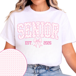 SENIOR pink varsity lettering with bow pattern fill and pink outline, EST. bow graphic 2026 printed on white short sleeve tee worn by female model, inset circle showing close up bow pattern detail