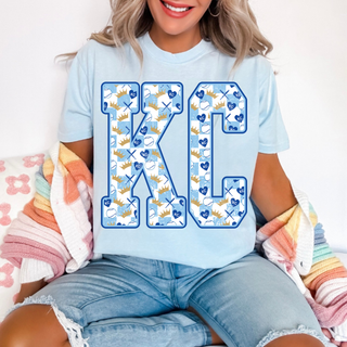 Woman wearing light blue KC crown baseball graphic shirt with large KC letters filled with crowns, baseballs, and heart icons in blue and gold.