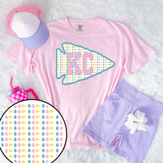 Pink Comfort Colors KC arrow graphic tee styled flat lay with pastel trucker hat, lavender shorts, and Easter egg pattern detail.