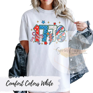 White t-shirt with colorful Red, White and Blue '1776' design worn by a person, with 'Seven Feathers Boutique & Custom Apparel' branding.