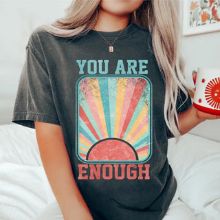 Dark charcoal You Are Enough Vintage Retro Graphic Top featuring distressed rainbow sunset graphic and bold affirmation text, worn by model holding a mug in a cozy indoor setting