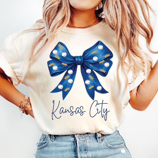 Woman wearing ivory Bella Canvas top featuring a blue baseball bow graphic with crown and baseball details and Kansas City script lettering.