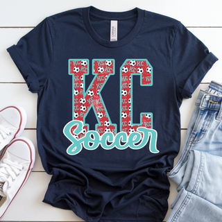Navy graphic top featuring large KC letters with red soccer print pattern and teal outline, layered with script Soccer across the front.