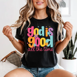 Bella Canvas black top featuring a colorful retro God Is Good All The Time graphic in bold pink, orange, teal, and yellow lettering with small floral accents, styled with denim shorts in a casual lifestyle setting.