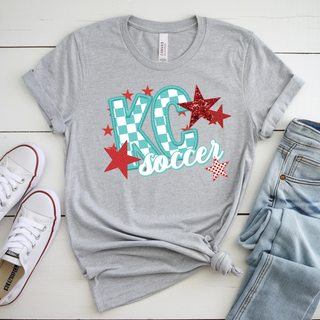 Heather grey Bella Canvas top featuring a KC soccer graphic with teal checkerboard KC lettering, bold red star accents, and white script soccer text layered across the front in a vibrant Kansas City inspired design.