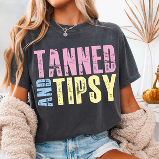 Woman wearing black Comfort Colors top with distressed Tanned and Tipsy graphic in pink, blue, and yellow lettering styled with denim shorts and layered jewelry.