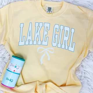 Comfort Colors butter yellow unisex Lake Girl graphic top featuring light blue varsity style lettering with a white bow underneath, styled in a flat lay with a Swig tumbler on a white textured background.