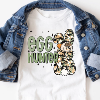 White graphic top featuring green “Egg Hunter” lettering with a camouflage Easter bunny wearing a hat, styled with a denim jacket.