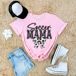 Soccer Mama faux glitter bow graphic top featuring distressed black varsity lettering with a large soccer ball bow design printed on a pink unisex garment styled in a flat lay with denim shorts and white sneakers.