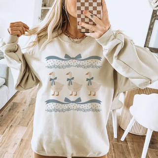Vintage three geese bow graphic top shown on model in sand featuring three white geese with blue bows, decorative ribbon accents, and soft farmhouse inspired detailing.