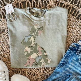 Folded sage green crewneck sweatshirt featuring a camouflage patchwork bunny graphic, styled with denim and accessories.