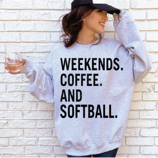 woman wearing grey sweatshirt with weekends coffee and softball graphic text holding drink and wearing black hat