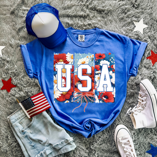 USA floral bow patriotic graphic top styled with denim shorts white high top sneakers blue trucker hat and american flag on neutral rug background