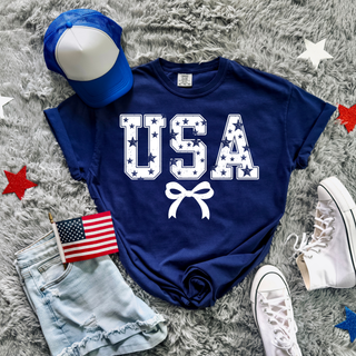 navy vintage usa stars graphic top with white star filled usa lettering and bow underneath styled with denim shorts, american flag, blue trucker hat, and white sneakers on grey blanket