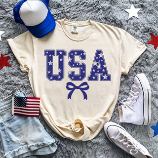 ivory vintage usa stars graphic top with blue star filled usa lettering and bow underneath styled with denim shorts, american flag, blue trucker hat, and white sneakers