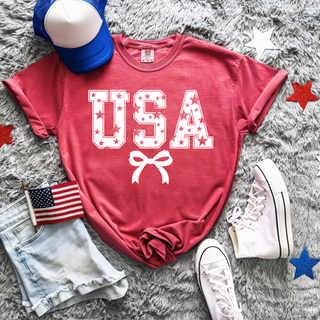 red vintage usa stars graphic top with white star filled usa lettering and bow underneath styled with denim shorts, american flag, and white sneakers
