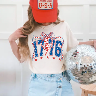woman wearing ivory 1776 patriotic graphic tee with blue star filled numbers and red bow detail paired with denim skirt and red trucker hat holding silver disco ball
