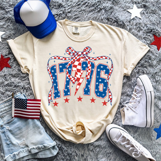 ivory 1776 patriotic graphic tee with blue star filled numbers, red checkered bow, and red stars styled with denim shorts, american flag, blue trucker hat, and white high top sneakers on grey textured blanket