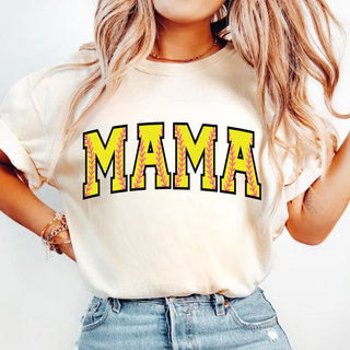 Woman wearing an ivory graphic top featuring bold yellow varsity-style “MAMA” lettering with pink softball stitch detailing inside each letter, styled casually with denim jeans in a neutral setting