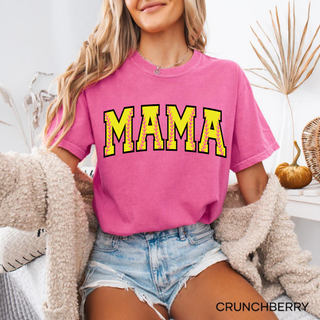 Woman wearing a bright pink crunchberry graphic top with yellow varsity “MAMA” text filled with pink softball stitch accents, styled with denim shorts and a cozy cardigan in a warm home setting