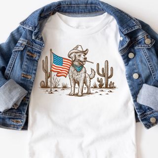 White graphic top styled with a denim jacket featuring a cowboy dog wearing a hat and bandana standing next to an American flag in a desert scene with cacti