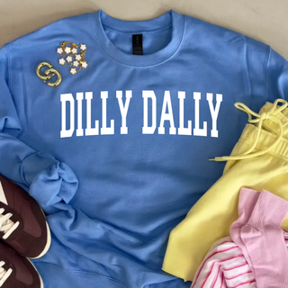 Blue graphic sweatshirt styled flat lay with bold white “Dilly Dally” text across the front, paired with yellow shorts, pink striped socks, and brown sneakers on a neutral background with accessories