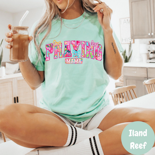 
Praying Mama Floral Bow Comfort Colors T-Shirt in Iland Reef, bright mint green tee with colorful tropical floral patterned letters and bow accents, worn by blonde woman holding iced coffee in kitchen setting