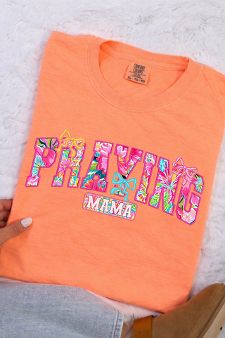 Praying Mama Floral Bow Comfort Colors T-Shirt in Neon Cantaloupe, folded orange tee with vibrant pink and teal floral patterned letters and bow graphic detail