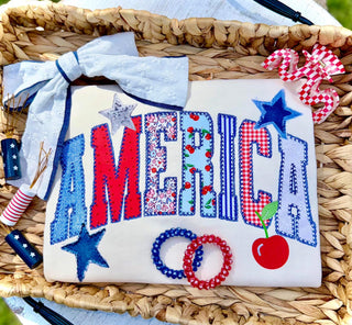 White tee with America patchwork faux embroidery design in red white and blue with stars and cherry, styled in wicker basket flat lay