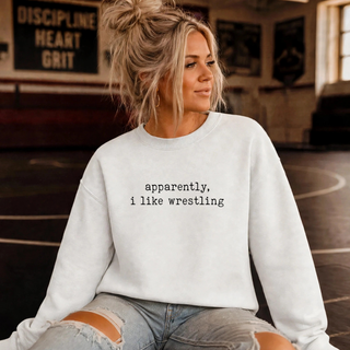 Woman in a white crewneck sweatshirt with black typewriter-style text reading apparently, i like wrestling, seated in a wrestling room