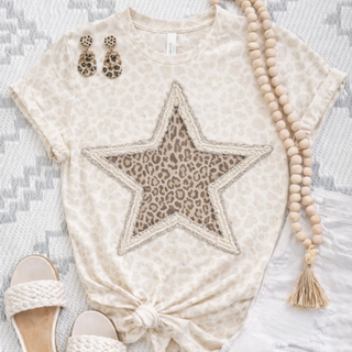 Neutral leopard print graphic top featuring a large frayed-edge star with layered leopard pattern detail, styled with white jeans, woven sandals, tassel necklace, and leopard earrings.