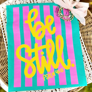 Island Green Comfort Colors tee with bold pink stripes and the words be still Psalm 46:10 in yellow script laid on a wicker basket with jewelry