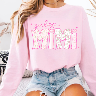 Woman in pink crewneck sweatshirt with blooming girly mimi floral bow graphic, denim shorts, sitting on a couch

