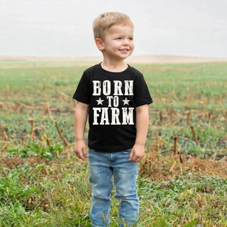 Toddler boy wearing black Born to Farm graphic tee with star design standing in a farm field