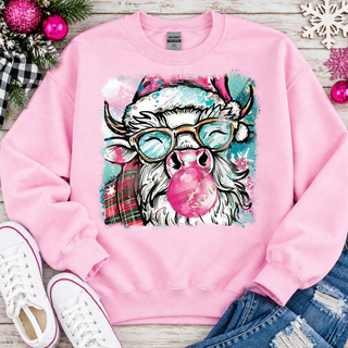 Light pink Gildan crewneck sweatshirt flat lay with a smiling Highland cow in a Santa hat and teal glasses blowing a pink bubblegum bubble, surrounded by pink ornaments, snowflakes, and white sneakers