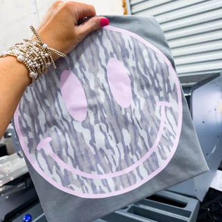Hand holding a grey Comfort Colors top with a large grey camo smiley face graphic with pink accents fresh off the DTF press