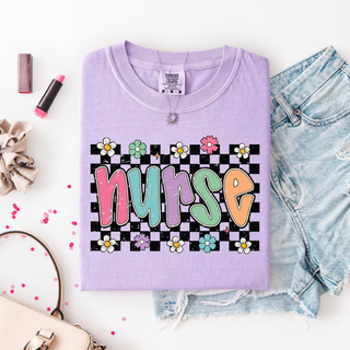 Folded violet Comfort Colors tee with nurse checkered daisy design, lipstick and denim flat lay
