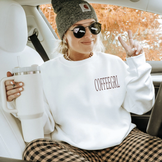 Woman in white crewneck with brown Coffeegrl left chest text holding a white tumbler in a car
