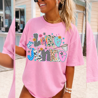 Woman wearing blossom Comfort Colors tee with colorful lake junkie patterned lettering, fish, daisies, and fishing net