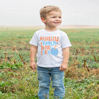 Toddler boy wearing white coolest pumpkin in the patch top with blue pumpkin and stars in a field