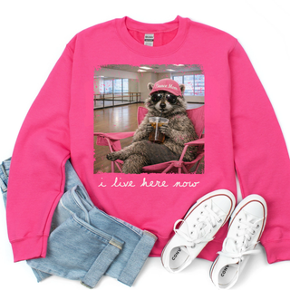 Heliconia pink Gildan crewneck sweatshirt with a raccoon wearing a Dance Mom hat and sunglasses sitting in a pink chair at a dance studio with the words i live here now
