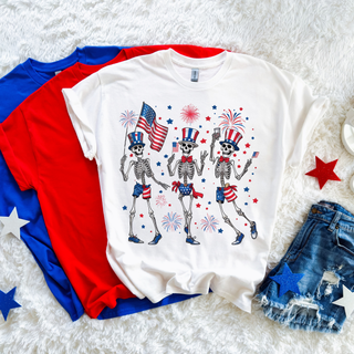White tee with dancing patriotic skeletons graphic featuring three skeletons in Uncle Sam hats and red white and blue outfits holding American flags with fireworks and stars