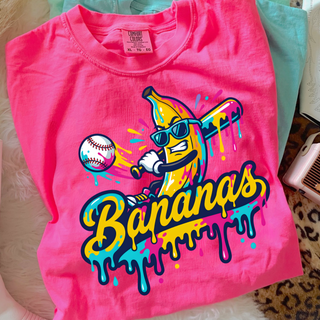 Hot pink Comfort Colors unisex t-shirt featuring a cartoon banana wearing sunglasses and holding a baseball bat, hitting a baseball. Colorful neon yellow, pink, and teal paint drip effect with the word “Bananas” in bold script.