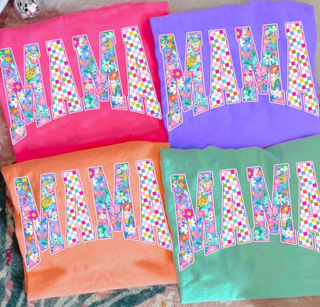 Comfort Colors short sleeve tees in crunchberry, neon pink, neon violet, and neon cantaloupe folded flat showing colorful floral and rainbow checkered MAMA block letter graphic